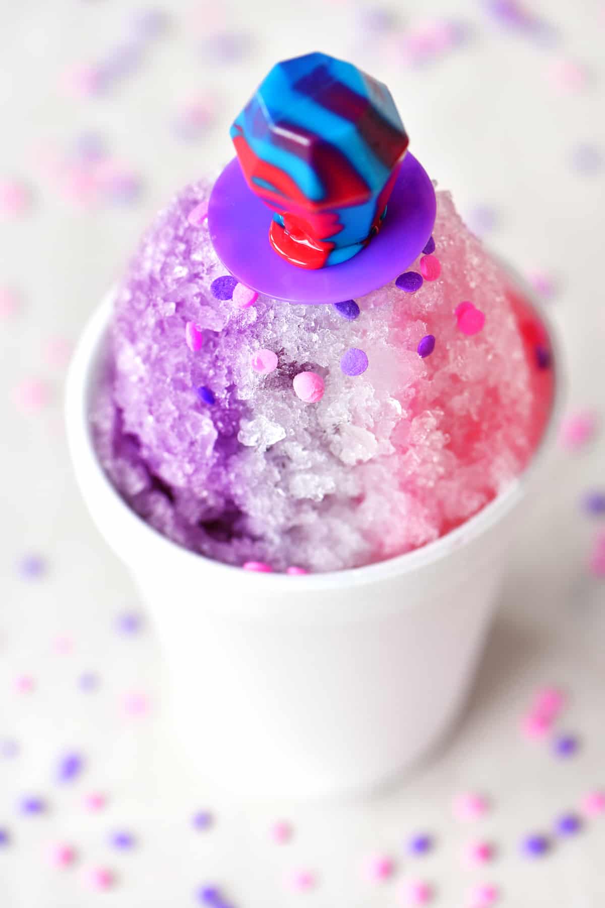a purple, white and pink themed shave ice treat with a ring-pop candy on top