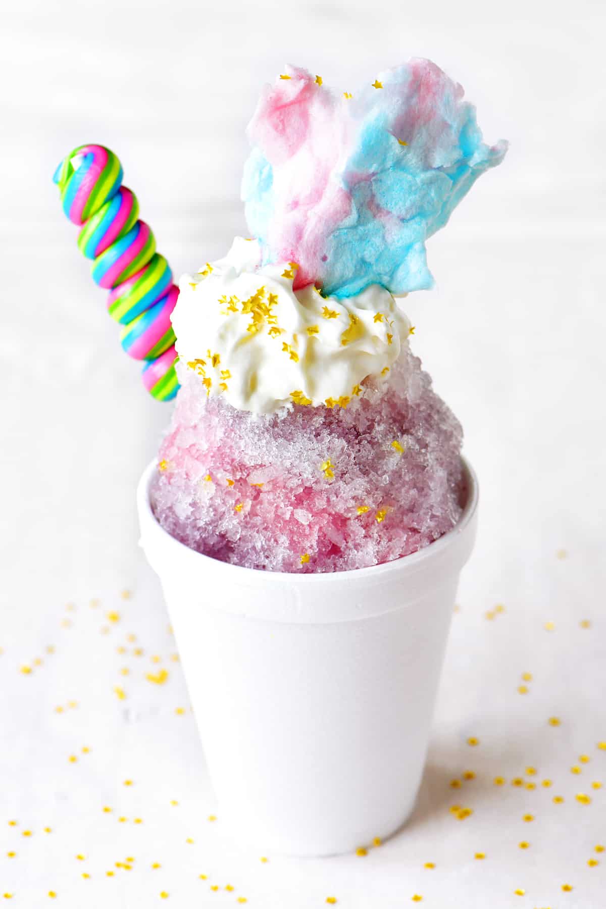 a unicorn themed pink and purple shave ice treat with a sucker and cotton candy on top