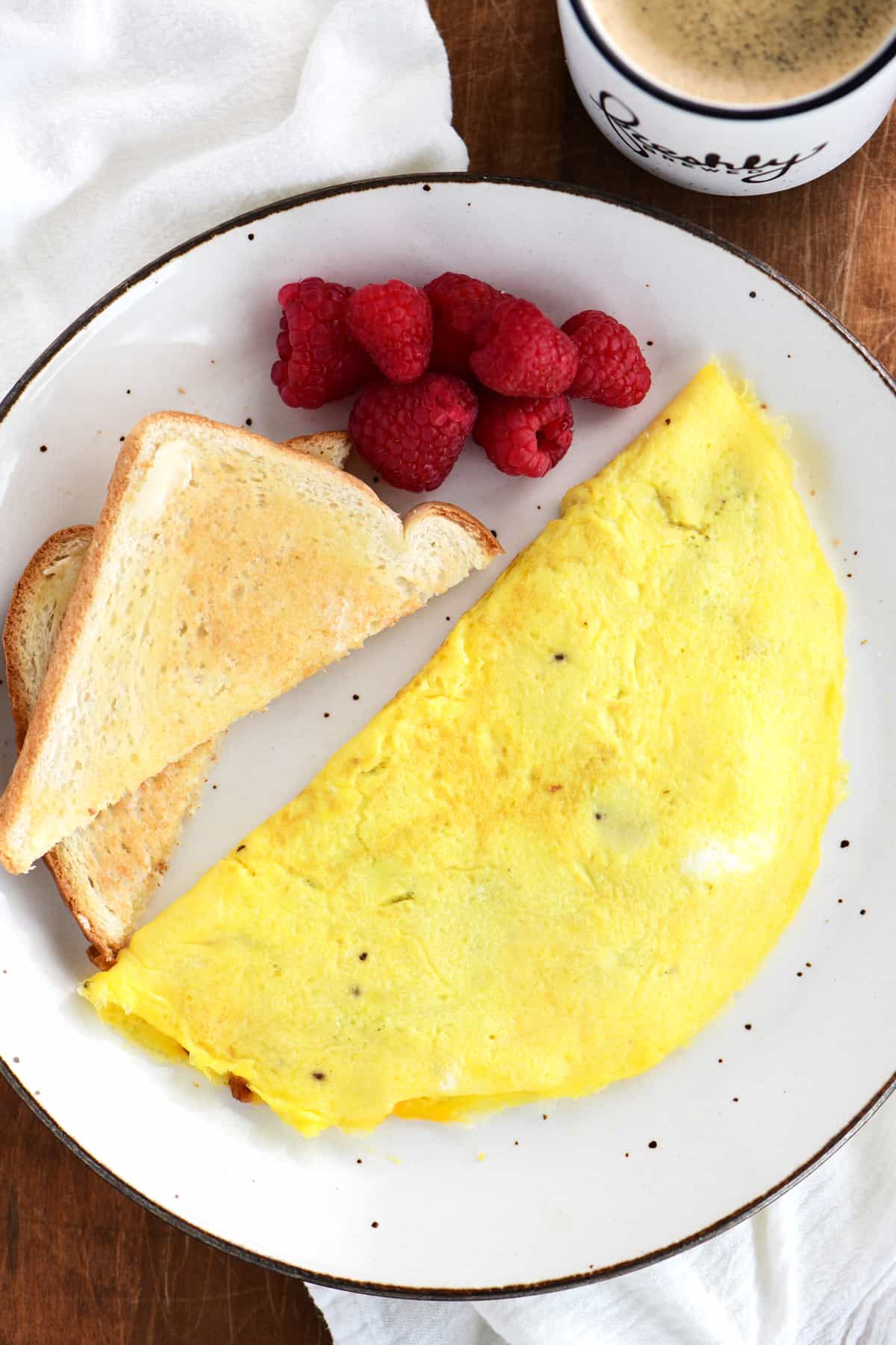 topdown view of an omelet, buttered toast and raspberries on a white speckled plate