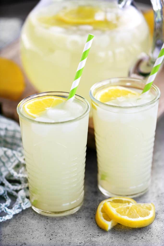 two glasses of lemonade with a pitcher in the behind them