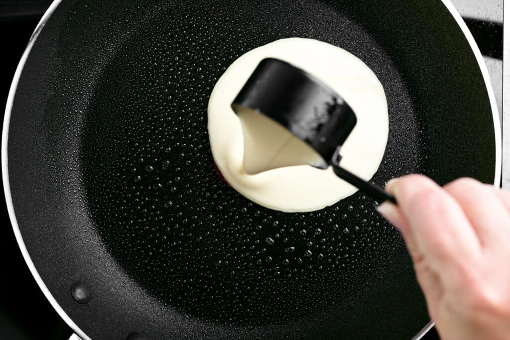 a hand pouring batter into a frying pan from a measuring cup