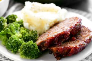 sliced meatloaf on a white plate with mashed potatoes and brococli