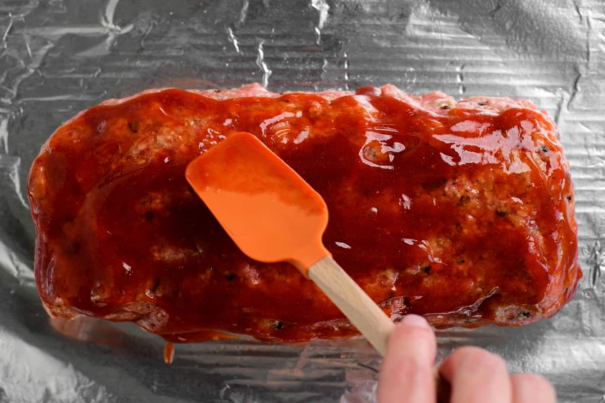 spreading glaze over meatloaf in pan with a orange spatula