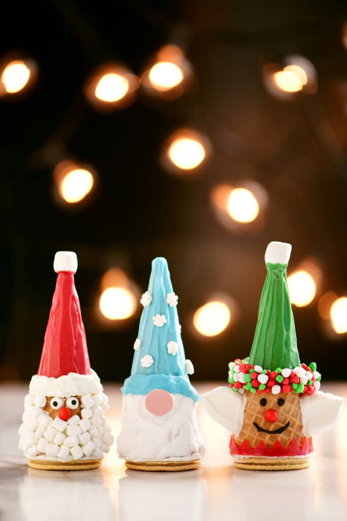 3 Christmas cones decorated as a Santa, Elf and Gnome on a table in front of holiday lites