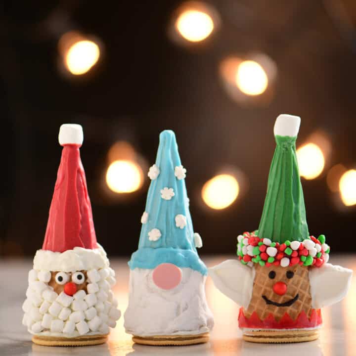 Christmas cones decorated as a Santa, Elf and Gnome set on a table in front of holiday lites