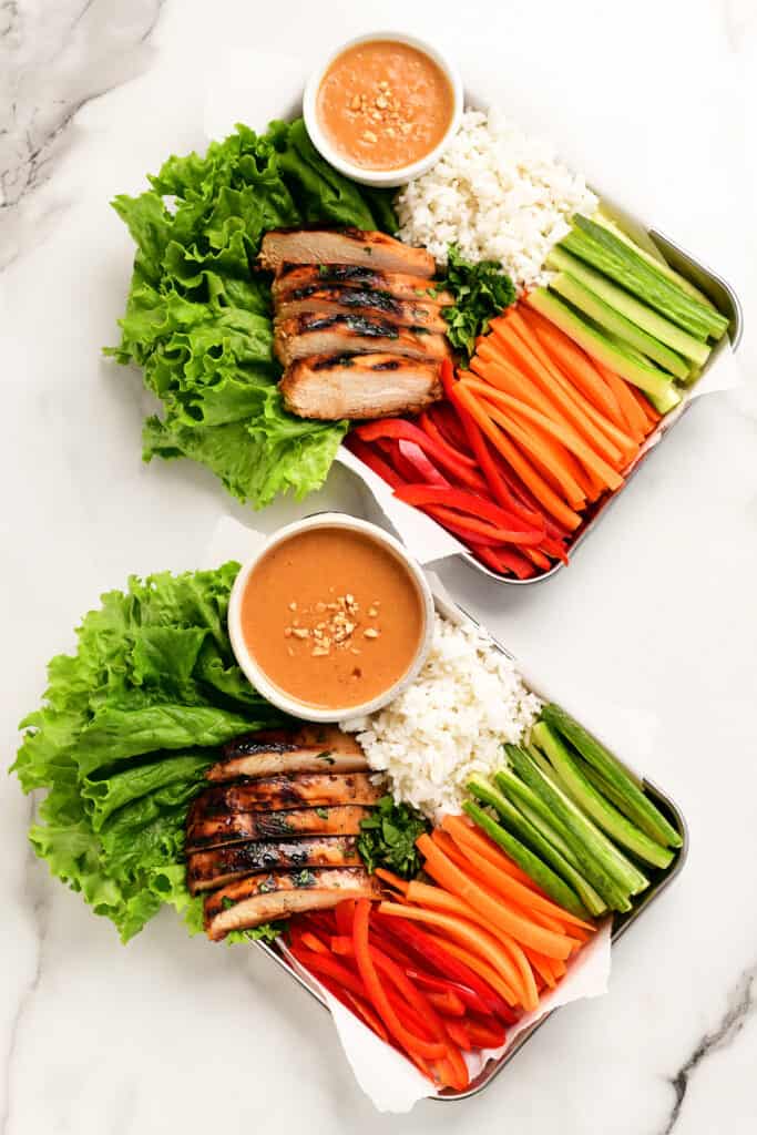two trays with sliced veggies, lettuce, rice, and chicken