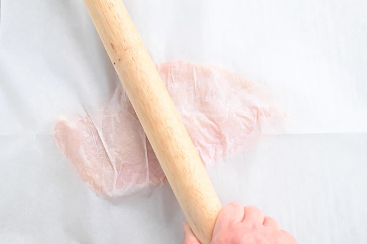 a hand using a wooden rolling pin to tenderize the uncooked chicken