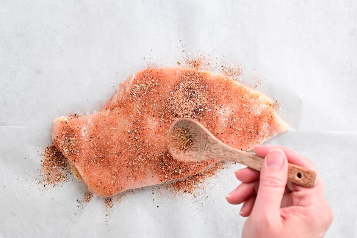 a hand using a wooden spoon to sprinkle spices on the uncooked chicken