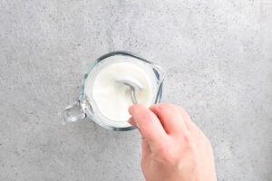 a hand using a spoon to stir buttermilk in a small glass pitcher