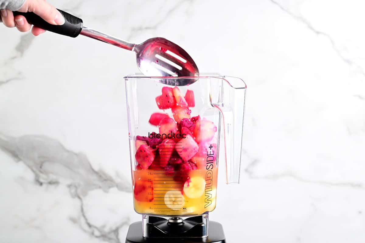a hand using a metal slotted spoon to add fruit to a blender