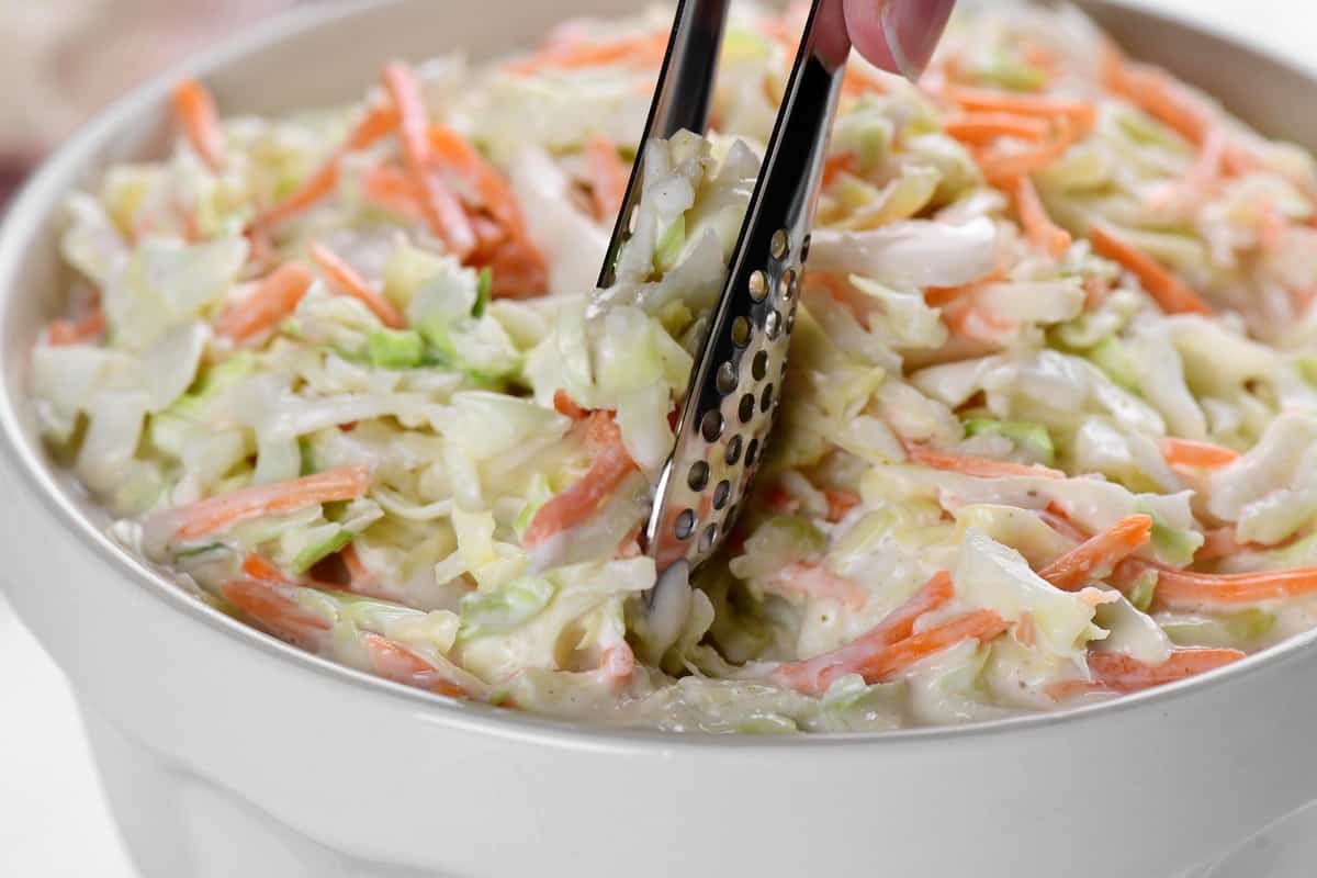 a hand using a pair of serving tongs to serve coleslaw from a bowl