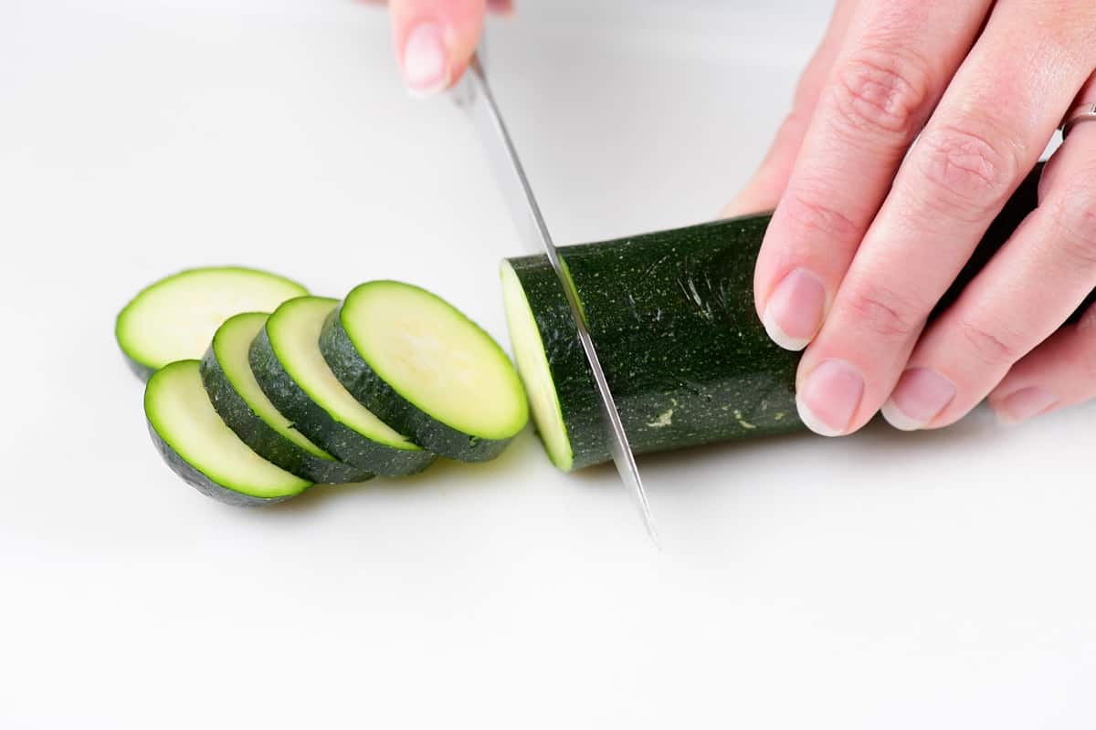 hands slicing a zucchini with a knife on a cutting board