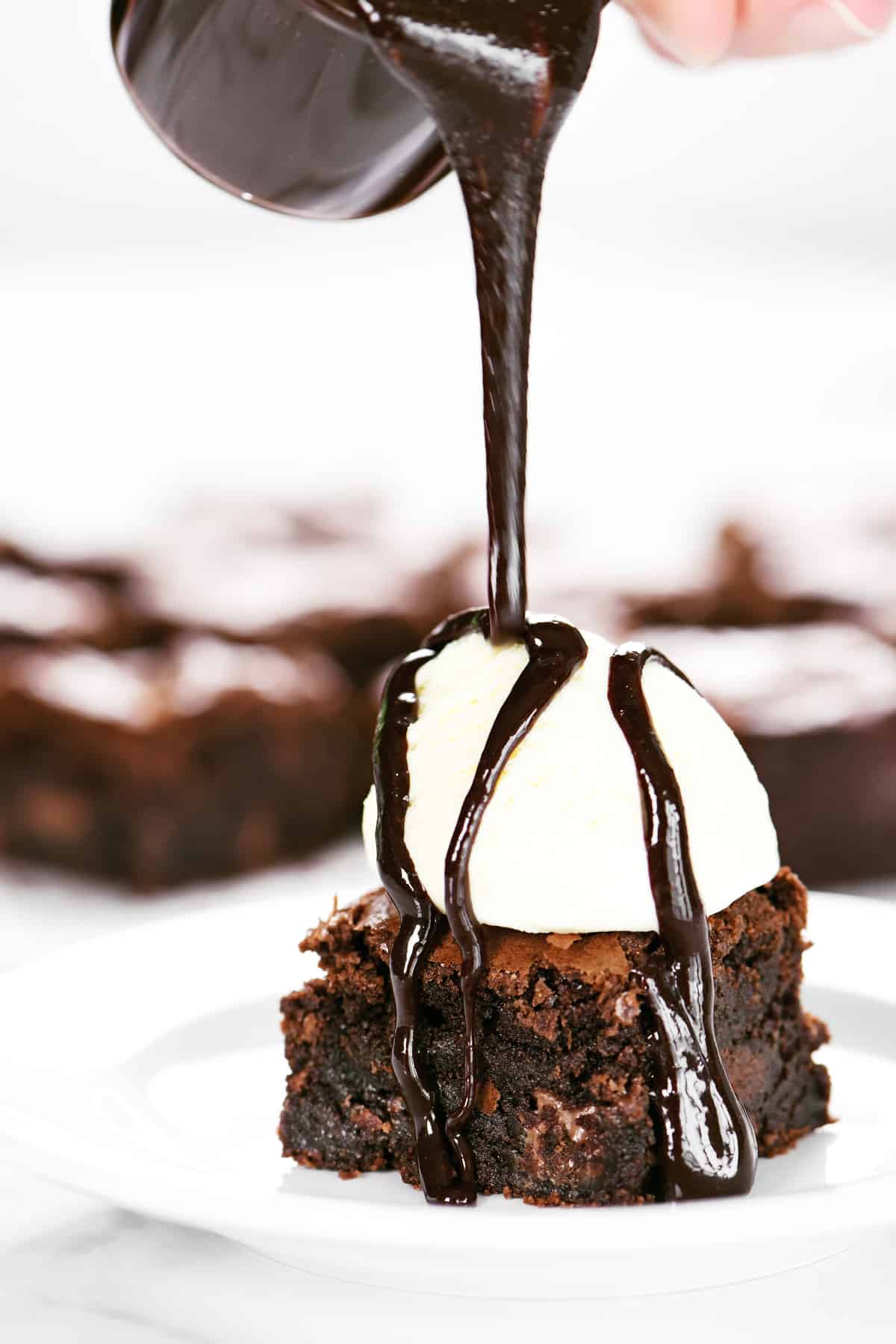 a hand pours hot chocolate sauce on to a brownie with vanilla ice cream.