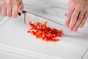hands using a kitchen knife to dice peppers on a cutting board.