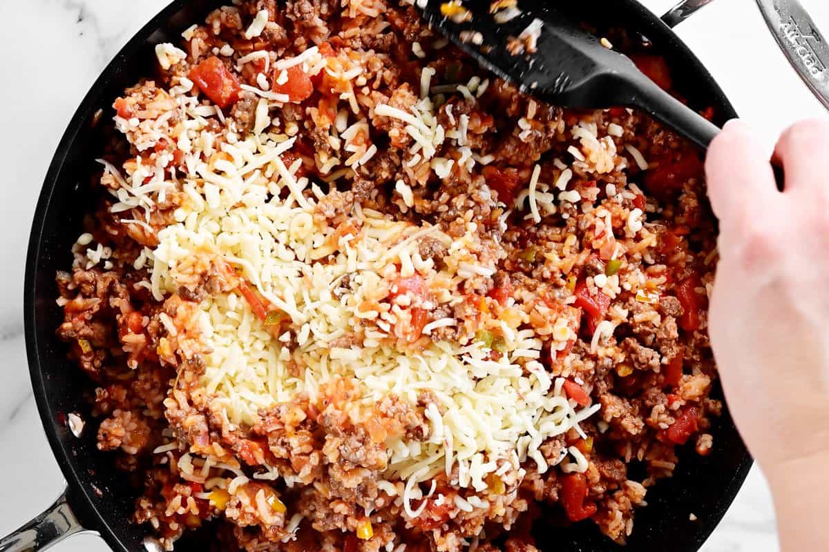 a hand using a black spatula to stir shredded cheese into the stuffed pepper ingredients.