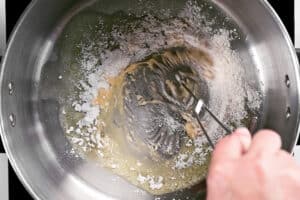 a hand uses a whisk to stir the roux ingredients.