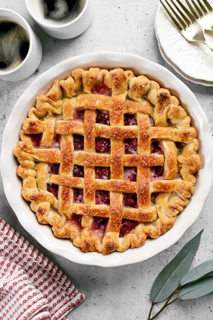 a cherry pie with a lattice top crust, two plates, two cups of coffee, and a red and white towel.