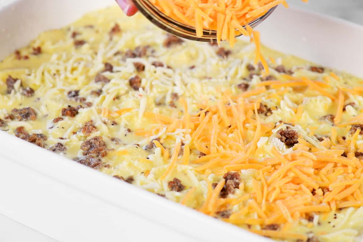 a hand pouring shredded cheddar cheese on to the uncooked casserole.