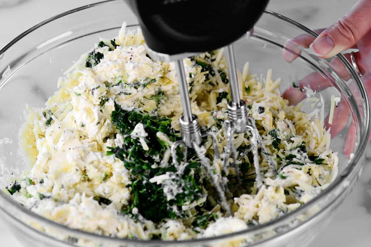 a hand holds a glass bowl while mixing spinach artichoke dip ingredients.