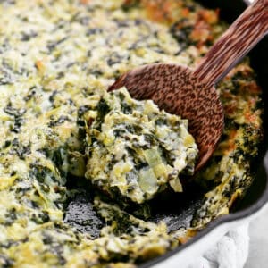 a wooden serving spoon scoops spinach artichoke dip out of the pan.