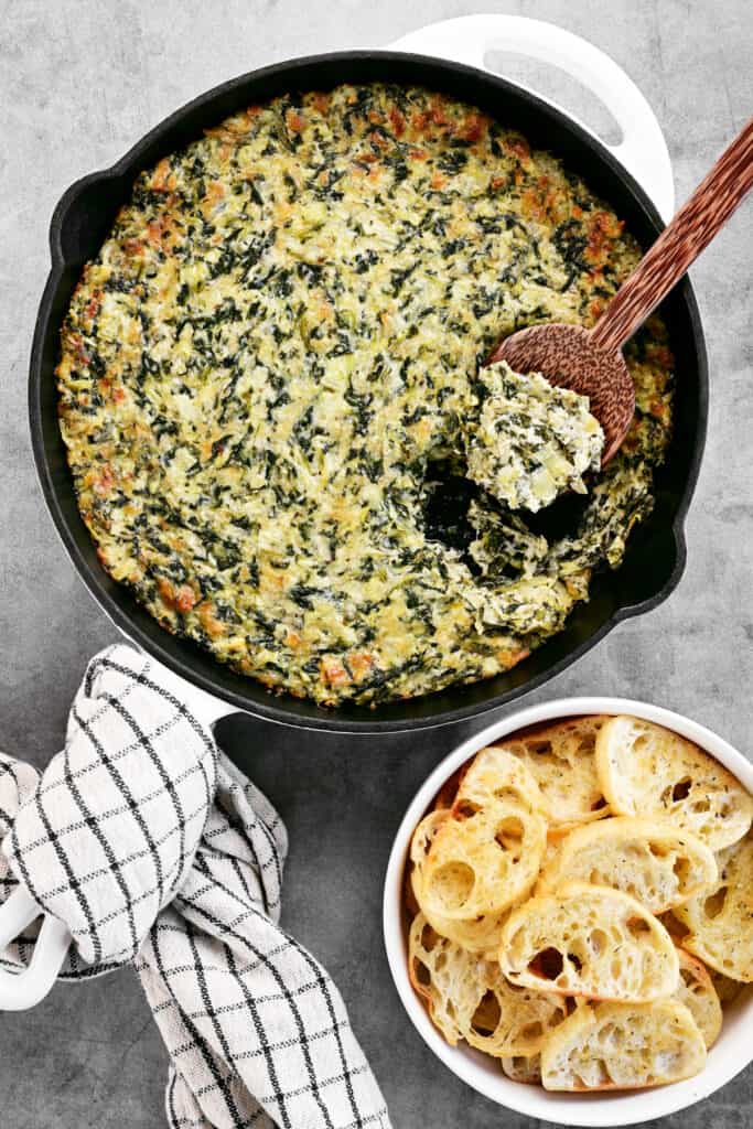 a pan of spinach artichoke dip with a bowl of crostini bread nearby.