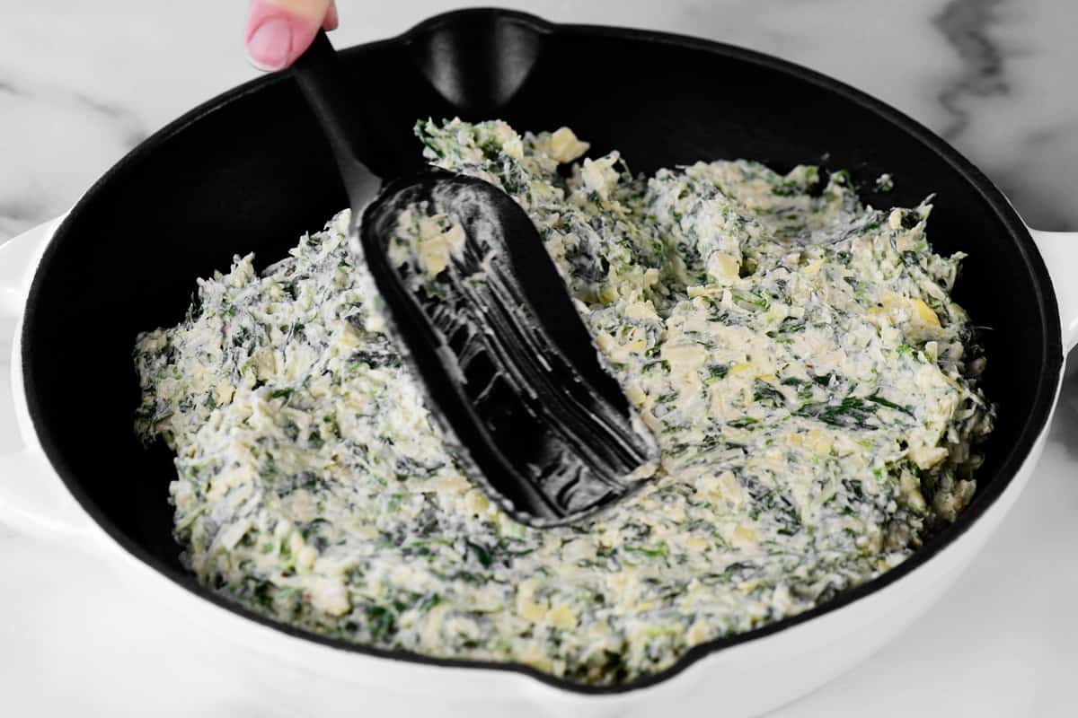 a hand using a rubber spoonula to spread the dip ingredients into an iron skillet.