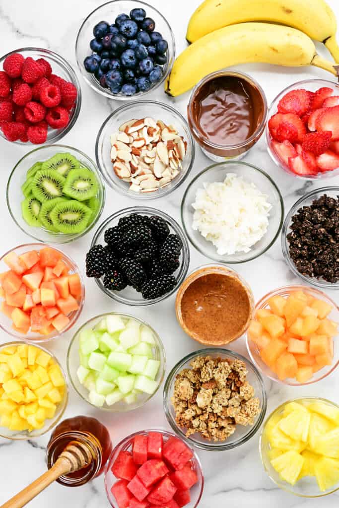 many containers with fresh fruit, granola, honey and acai bowl toppings on a countertop.