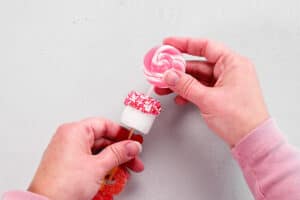 hands sliding a lolli pop into a marshmallow that is on top of the candy stick.