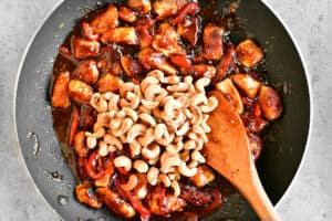 a wooden spoon stirs cashews, chicken and sauce in a frying pan.