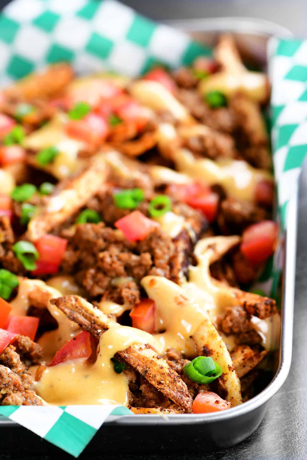 A silver tray with green checkered liner topped with crispy potato wedges, taco meat, tomatoes, and cheese sauce.
