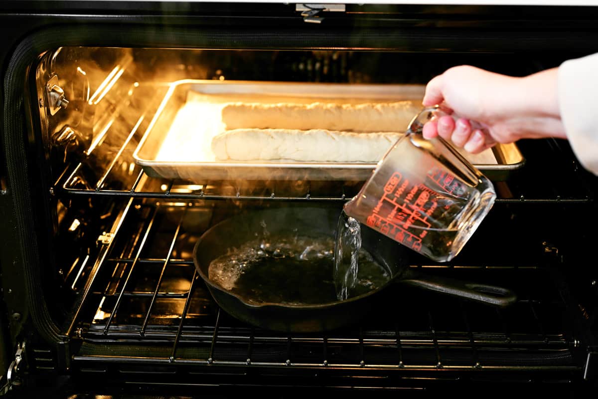 a hand pouring water from a glass pitcher into a hot iron pan.