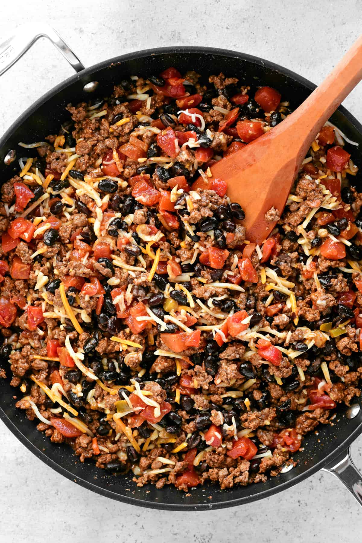 meat, tomatoes, beans, and cheese in a skillet with a wooden spoon.