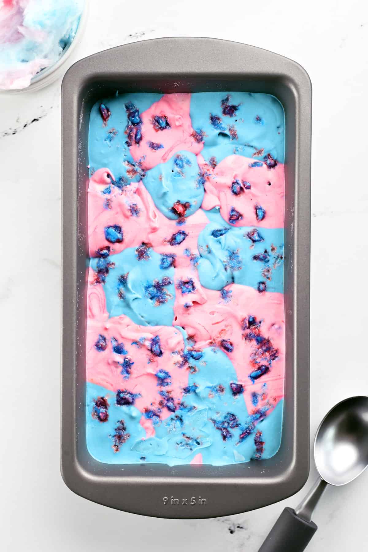 Cotton candy ice cream in a pan with an ice cream server and a bowl of cotton candy set nearby.