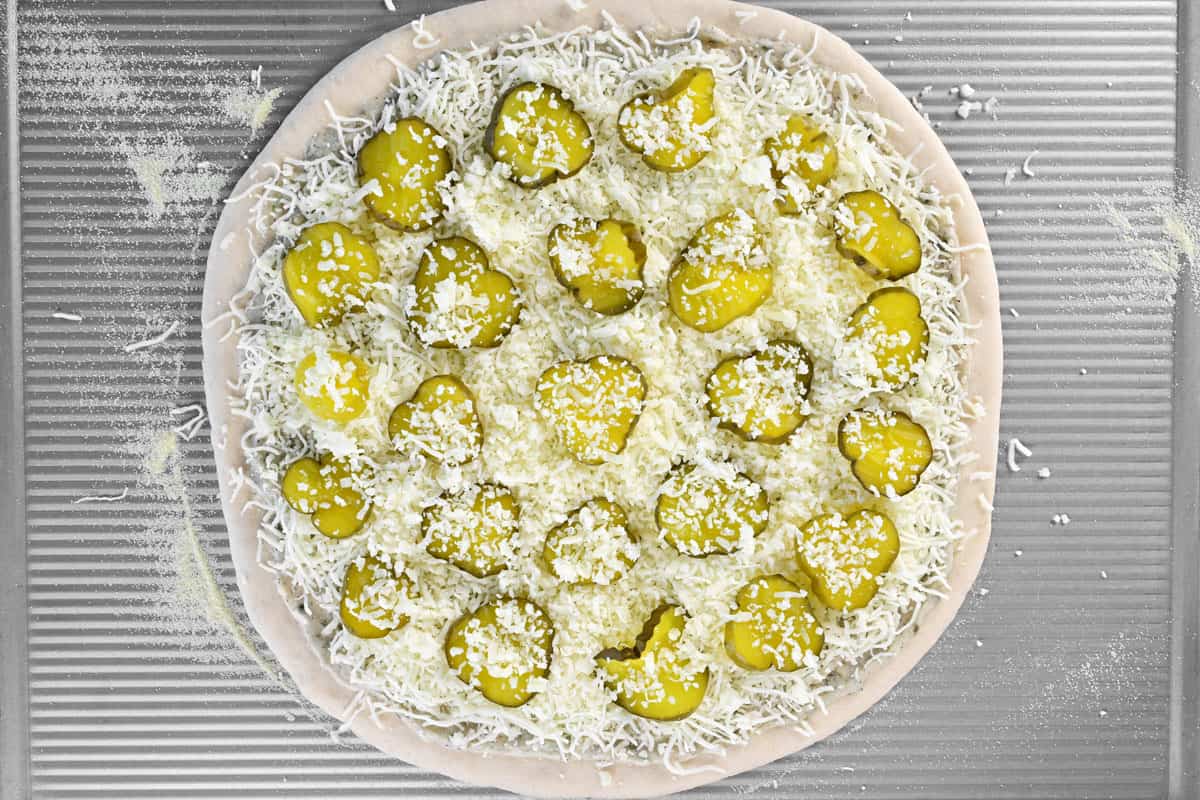 A pizza dough with pickles, sauce, and cheeses on it.
