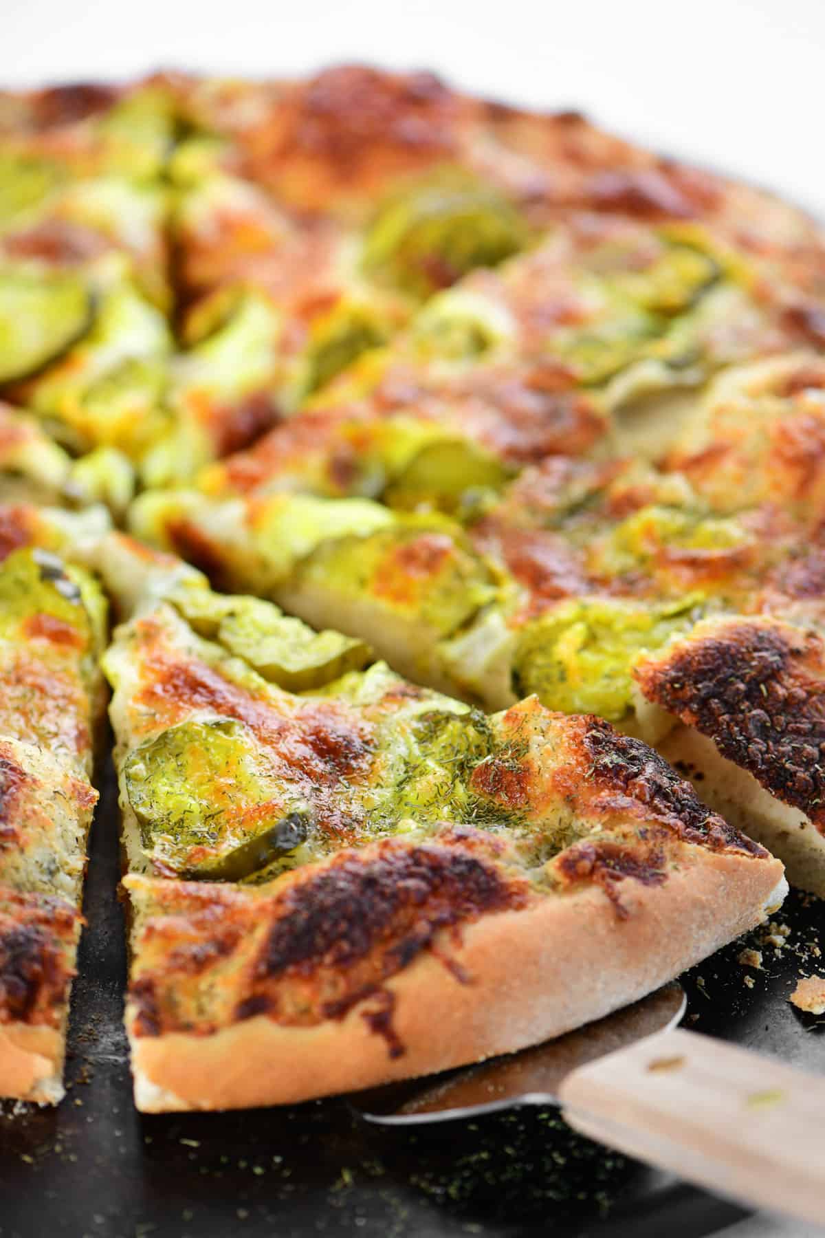 A serving utensil is shown underneath a slice of pickle pizza.