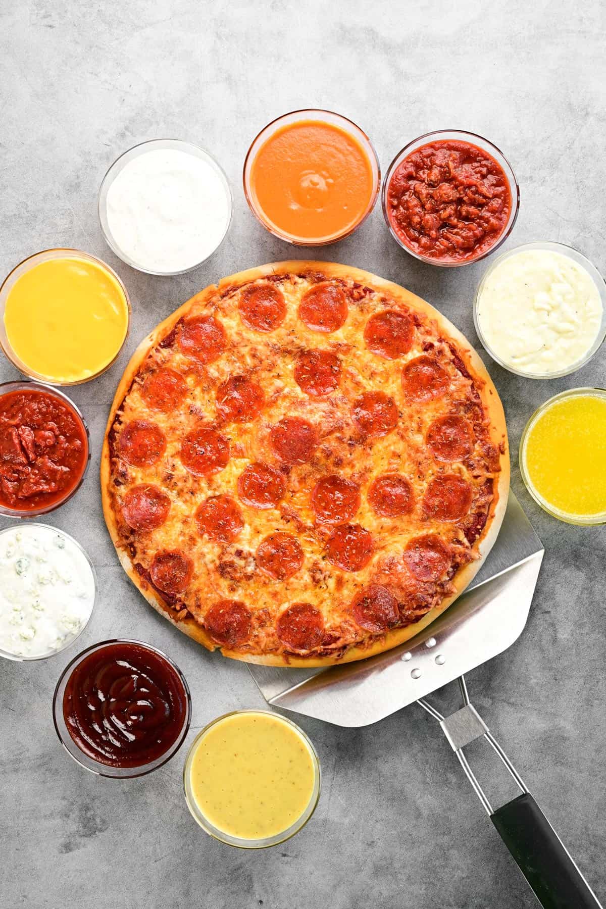 A pizza on a pizza peel with bowls of various dipping sauces around it.