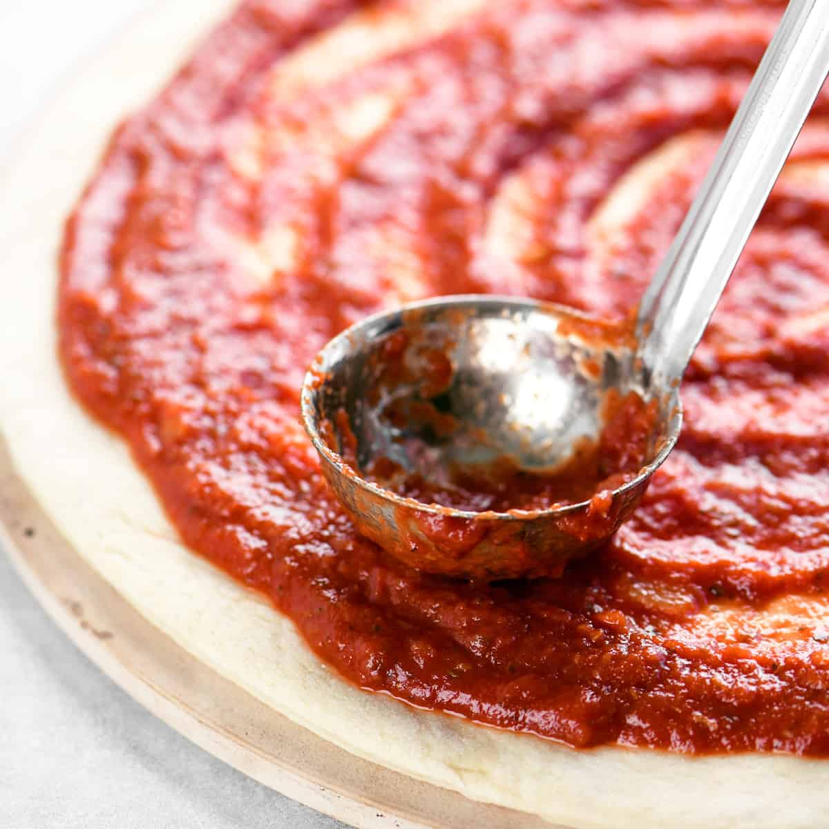 A ladle resting on pizza dough with sauce on top.