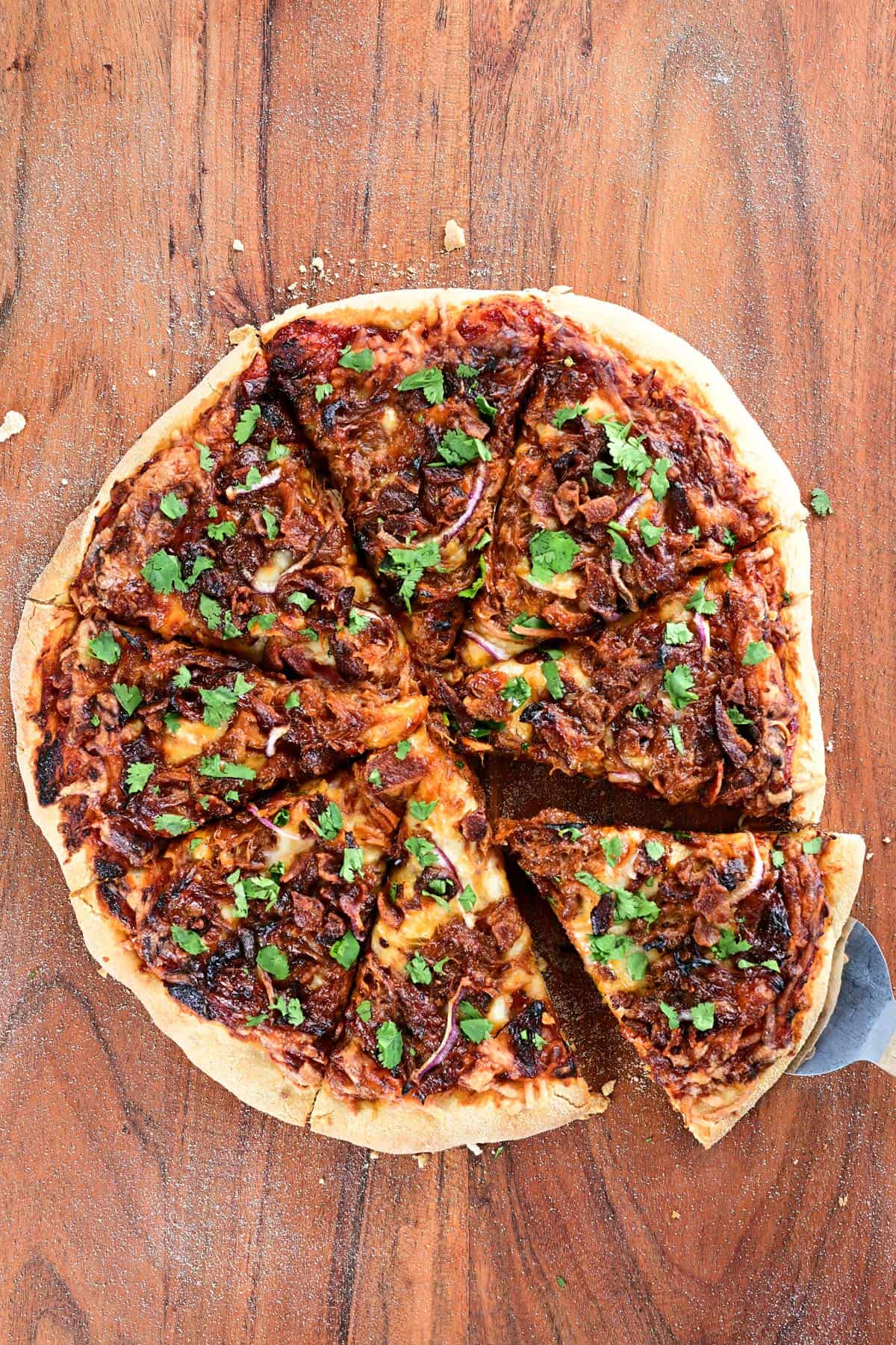 A slice of pizza is removed from a pulled pork pizza that is resting on a wooden cutting board.