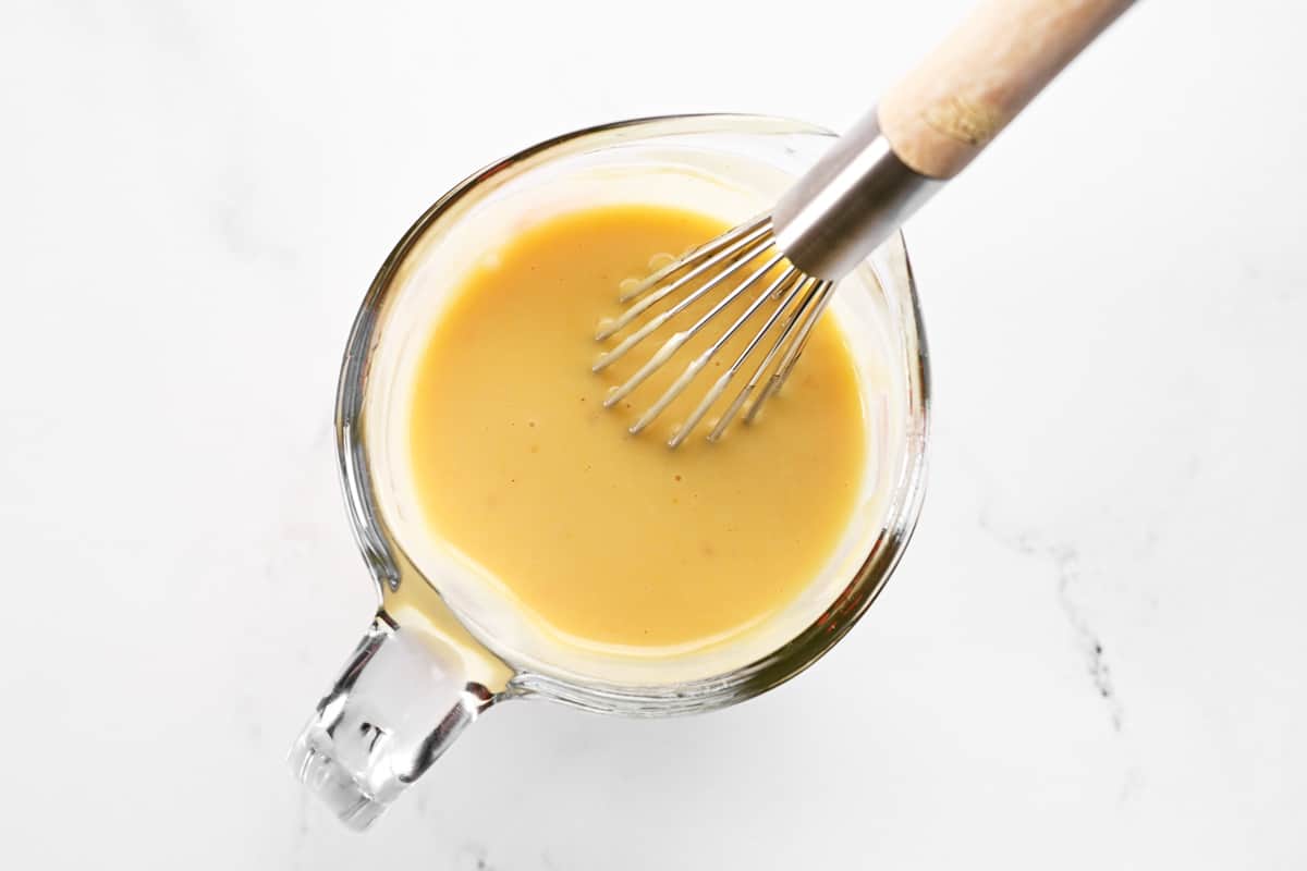 A whisk and mixed mustard sauce in a glass measuring cup.