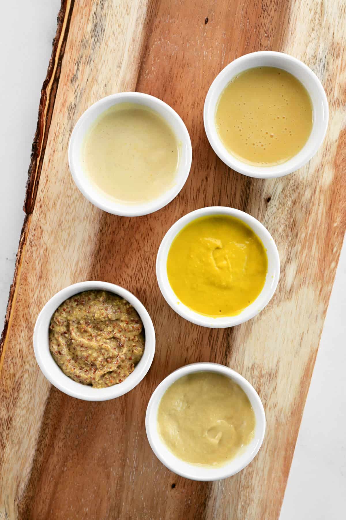 Five Dijon mustard substitutes in small bowls on a wooden cutting board.