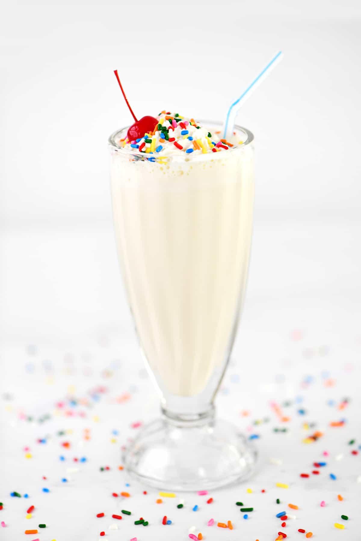 A vanilla milkshake rests on a countertop with candy sprinkles all around it.