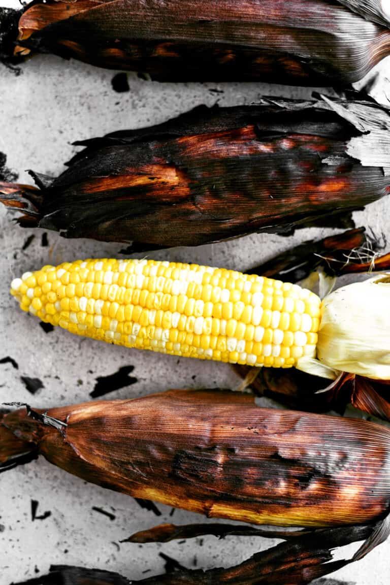 Three grilled cobs of corn in blackened husks with one peeled cob of corn on a pan.