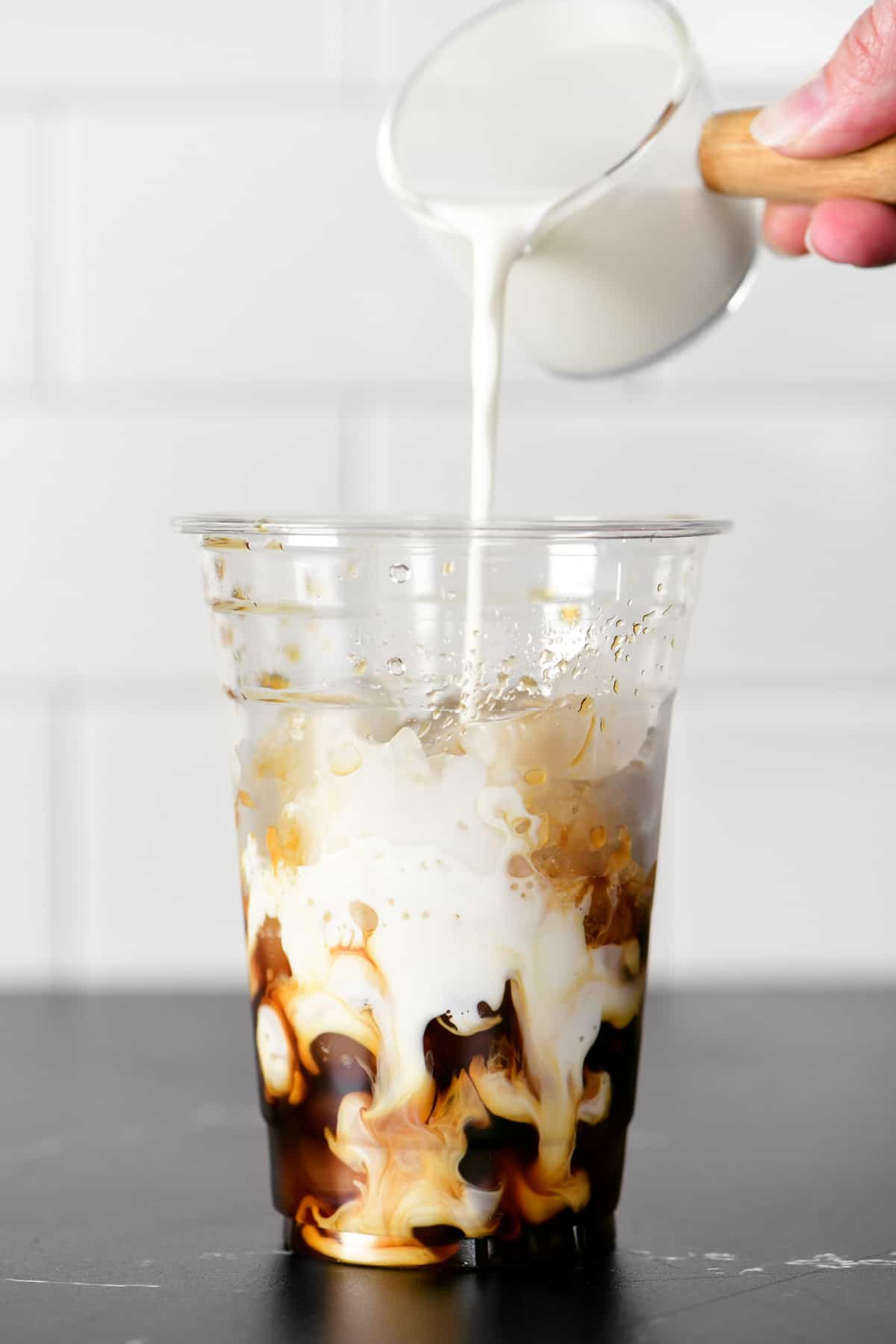 A hand pouring cream into a cup of iced coffee.
