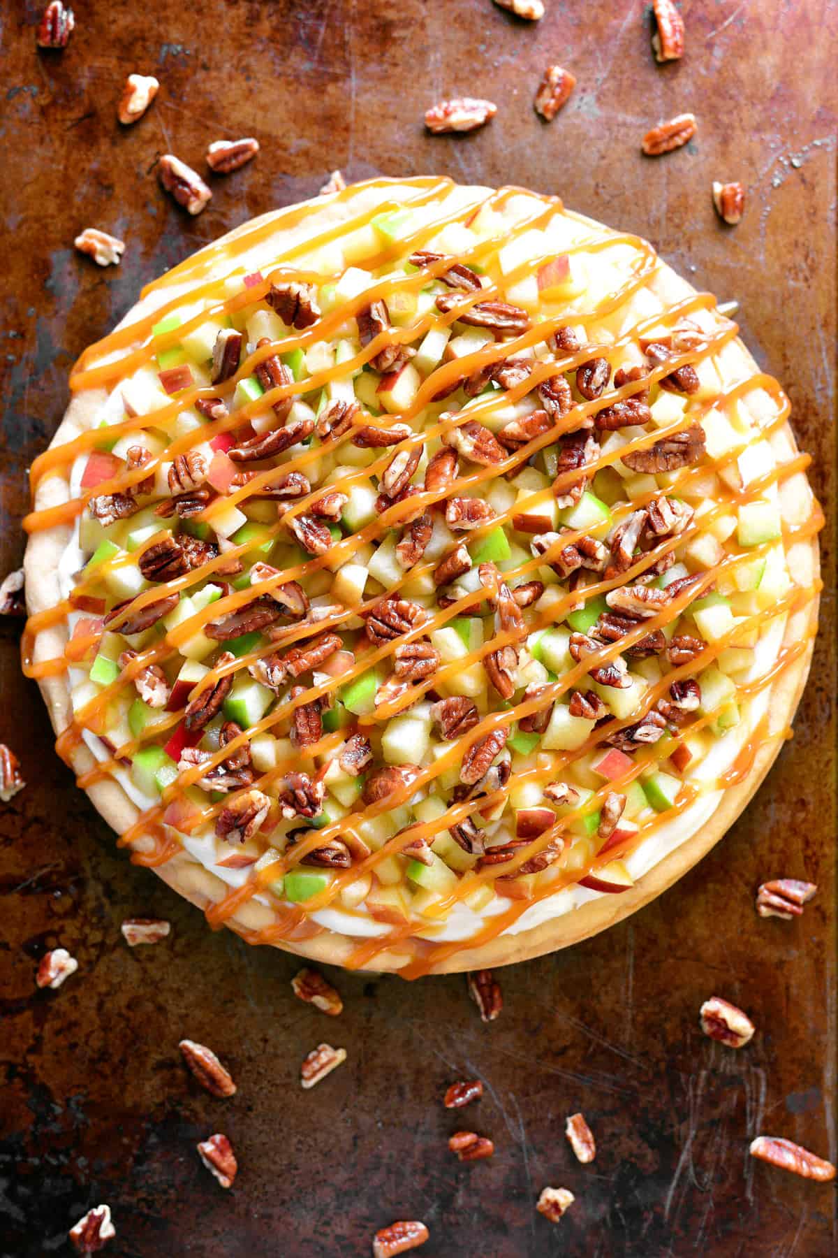 A caramel apple fruit dessert pizza on a pan with pecans sprinkled around it.