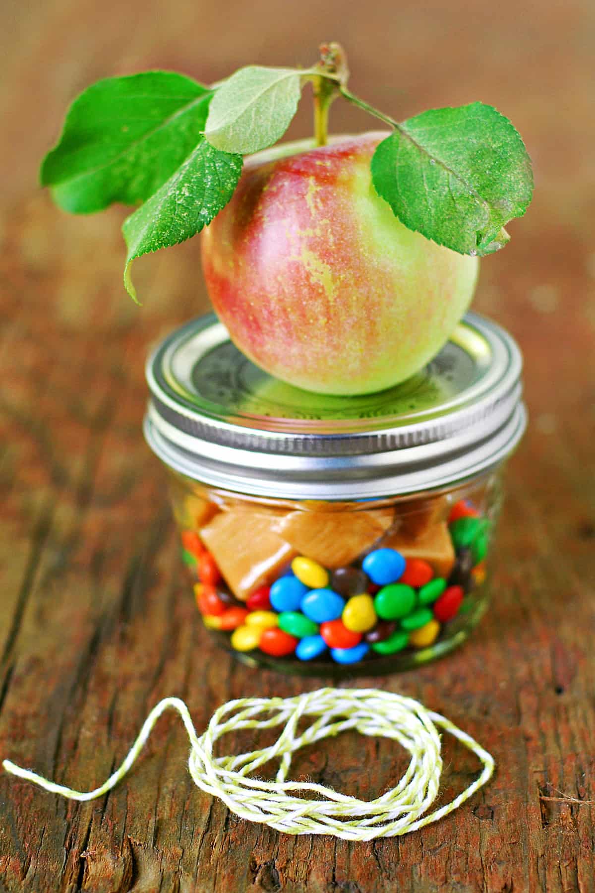 An apple with leaves attached sitting on top of a glass jar with candy inside.