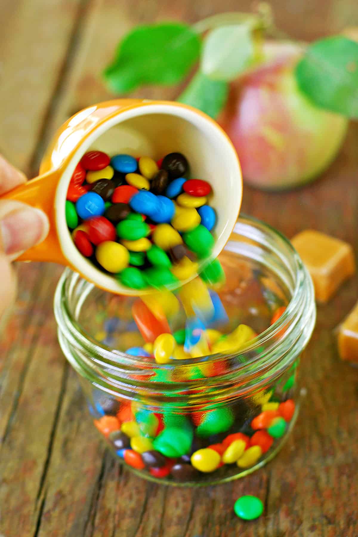 A hand is pouring candy from a measuring cup into a glass jar.