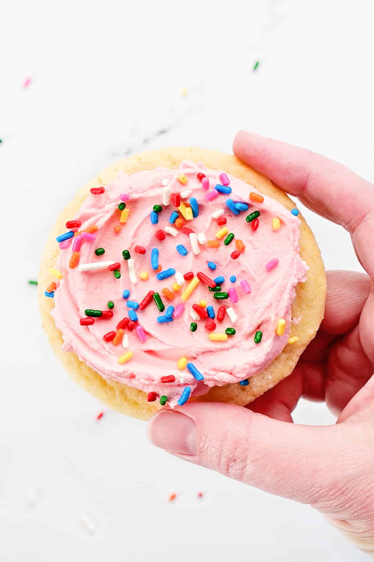 A hand holding a sugar cookie with pink buttercream frosting and sprinkles.