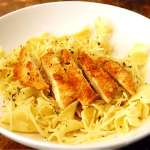 Sliced parmesan chicken on buttered noodles in a white bowl.