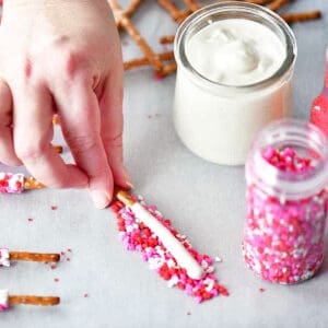 A hand rolling a candy coated pretzel in pink and red sprinkles.