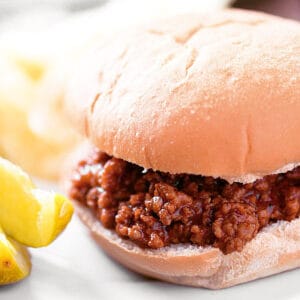 Sloppy joes on a white plate with pickles and chips.